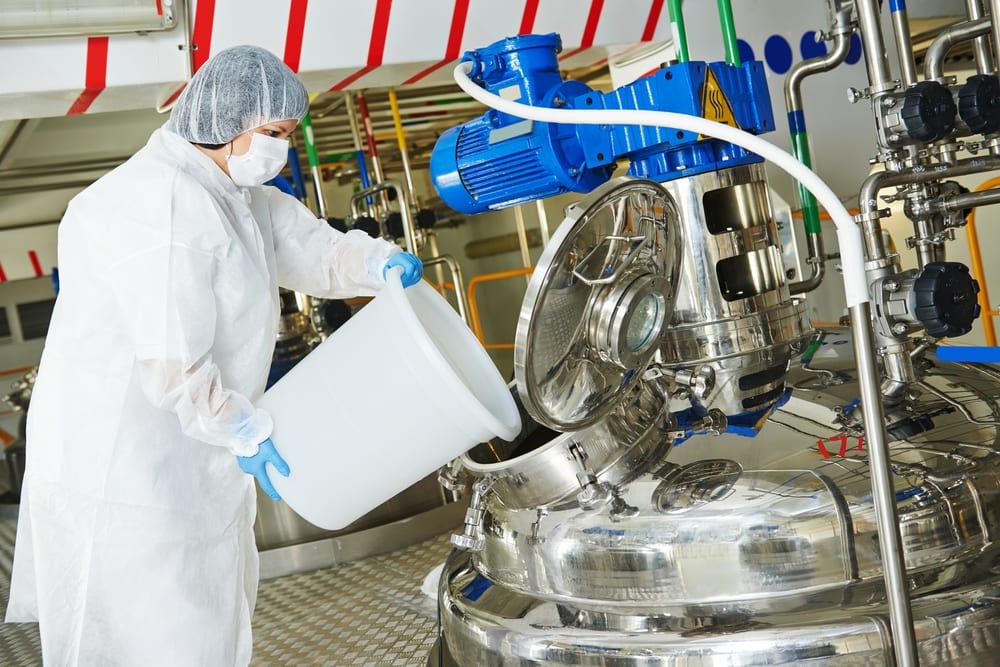 A technician in protective gear adding ingredients to a manufacturing mixing vessel
